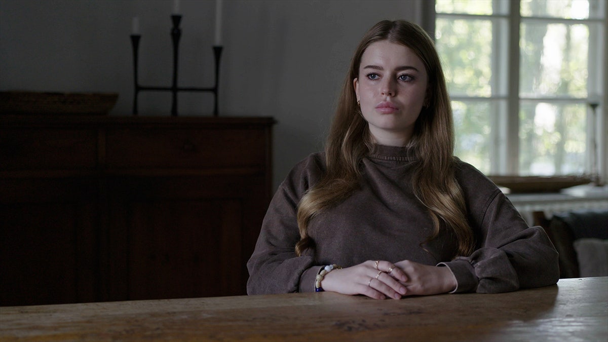 Jamie-Lee Arrow wearing a dark brown sweater sitting in front of a wooden table.