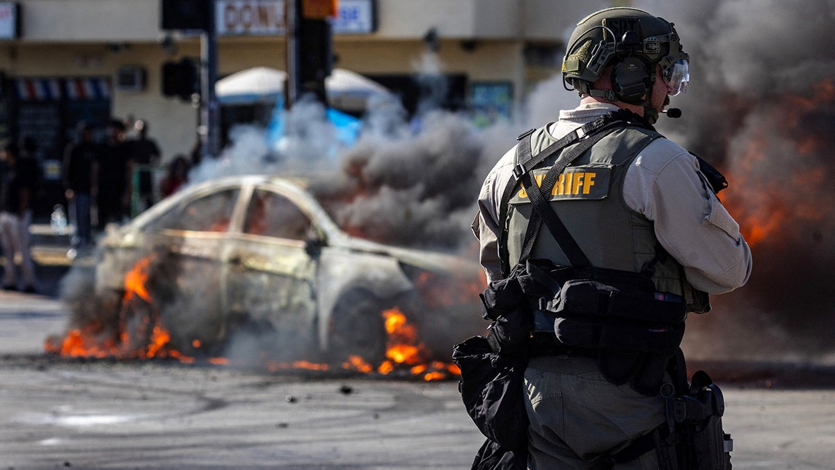 A car on fire with a law enforcement office looking on at protest