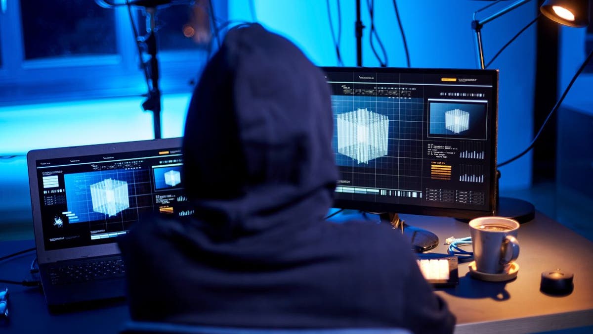 Person wearing a hoodie works on multiple computer screens displaying digital data in a dark room.