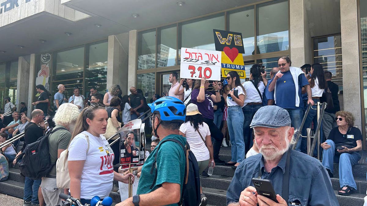 Crowd gathers on steps in Tel Aviv holding pro-Trump and protest signs during daytime event.