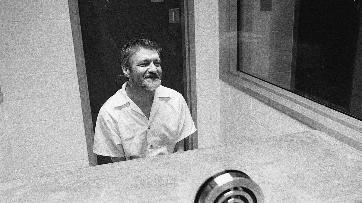 A black-and-white photo shows Ted Kaczynski seated in a prison visitation room wearing a light-colored short-sleeve shirt.