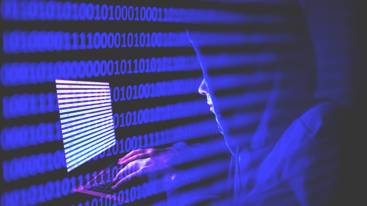 A person is seen working on a laptop that shows rows of binary code.
